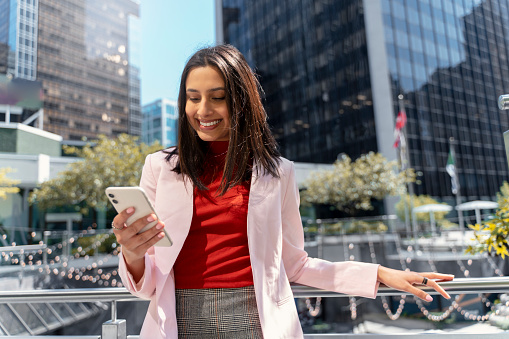 Beautiful smiling Indian woman holding mobile phone reading text message standing on urban street. Happy asian female using mobile app shopping online outdoors. Mobile banking, technology concept