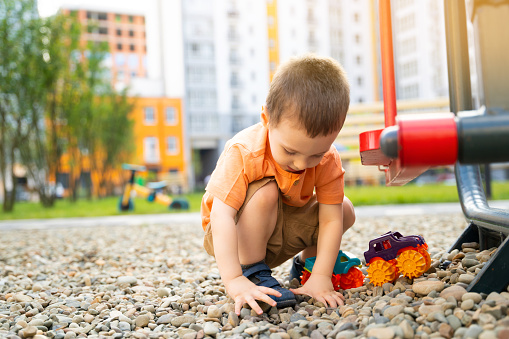 Little toddler boy playing with cars on pebbles on the playground. Boy two or three years old. Toddlerhood childhood concept. Residential complex of apartment buildings.