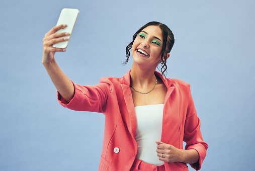 Phone, selfie and woman with a smile from social media and profile picture for creative job in studio. Influencer, happy and female person with makeup and fashion with blue background and model