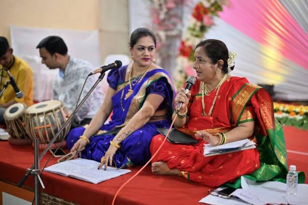 In this enchanting scene, two beautiful Indian women, draped in exquisite traditional attire, sit gracefully on the stage, their voices weaving a melodic tapestry as they sing a classic song accompanied by a skilled Tabla musician