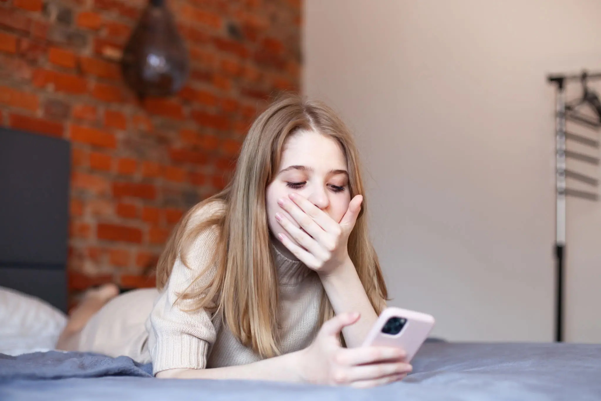Surprised teen girl lying on bed looking at smartphone covering mouth with hand.