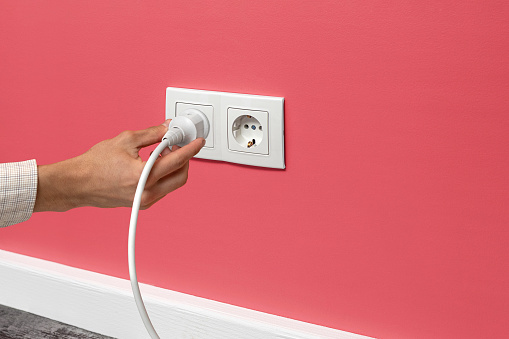 Human hand holding white cable plugged into double white electrical outlet on the pink wall