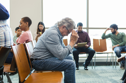 The senior adult man sits anxiously in the waiting room with his hands against his forehead.