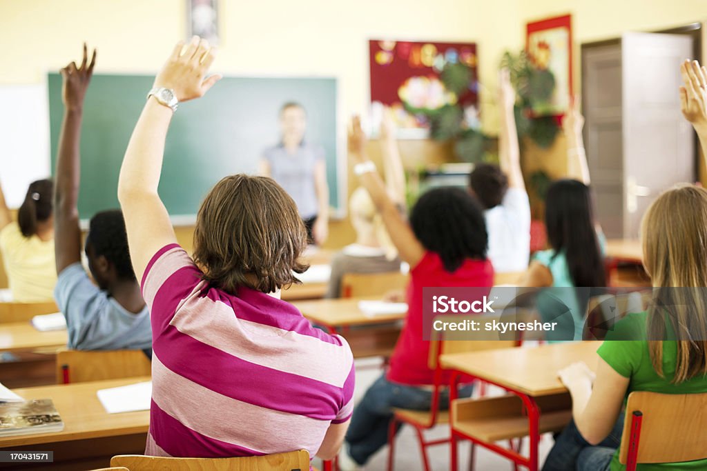 Group of teenagers sitting in the classroom with raised hands. Rear view of the teens sitting in the classroom and raising hands to answer on the question.   Rear View Stock Photo