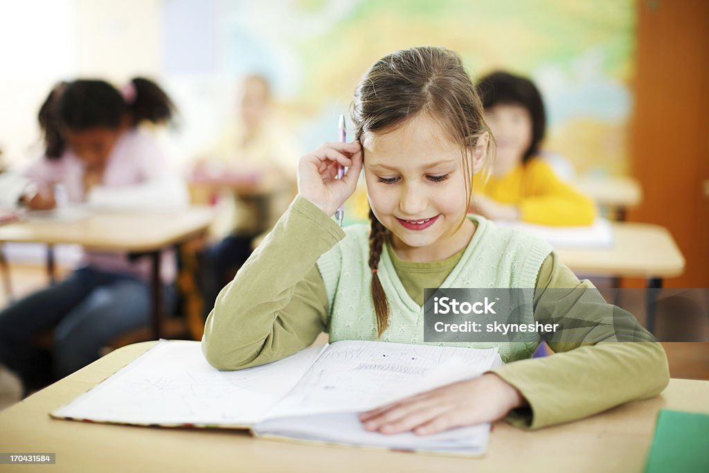 Smiling girl sitting in the classroom Beautiful schoolgirl sitting at the classroom. In the background is a group of pupils.   Book Stock Photo