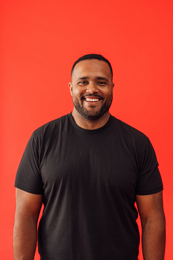 A portrait against a red background in a studio. He is looking confidently into the camera and is wearing a black tee shirt.