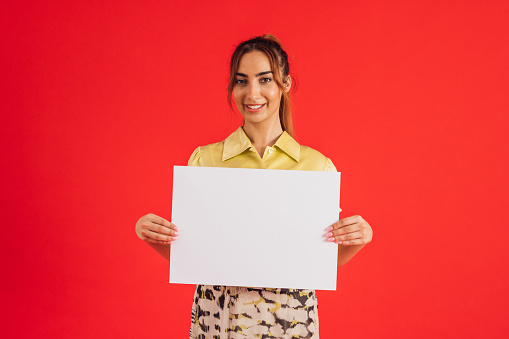 A close up red background studio shot of a young woman smiling and looking into the camera with a content look on her face. She is holding a white sign which is blank.