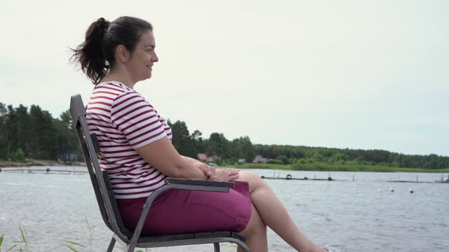 a middle-aged woman is sitting on a chair on the shore of a lake enjoying