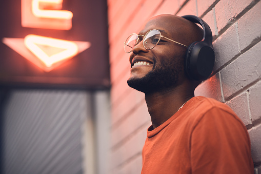Thinking, smile and music with a black man in the city, leaning on a brick wall on the street at night. Idea, glasses and headphones with a happy young male person streaming or listening to audio