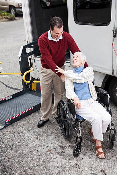 Senior woman by minibus with wheelchair lift Man helping senior woman (70s) in wheelchair, beside shuttle bus equipped with wheelchair lift. wheelchair lift stock pictures, royalty-free photos & images