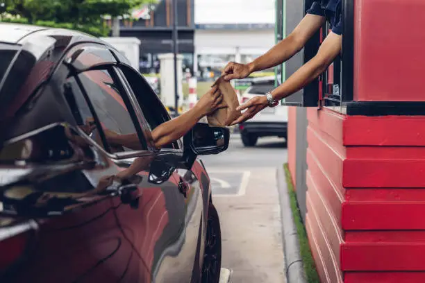 Photo of Hand Man in car receiving coffee in drive thru fast food restaurant. Staff serving takeaway order for driver in delivery window. Drive through and takeaway for buy fast food for protect covid19.