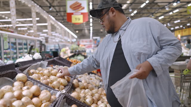 Fat man selecting onion from the produce stand and putting them in plastic bag while shopping in a supermarket for home cooking on weekend.