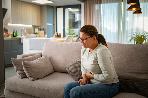 Woman suffering heavy stomachache sitting on the couch. High resolution 42Mp indoors digital capture taken with SONY A7rII and Zeiss Batis 40mm F2.0 CF lens