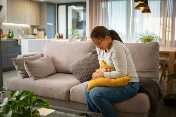 Unhealthy mature woman with stomachache sitting on the couch Woman suffering heavy stomachache sitting on the couch. High resolution 42Mp indoors digital capture taken with SONY A7rII and Zeiss Batis 40mm F2.0 CF lens gastroenteritis stock pictures, royalty-free photos & images