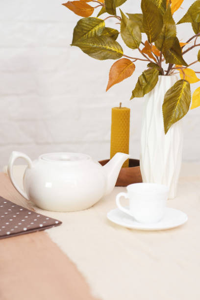 Autumn home decoration. Vase with autumn golden leaves branches, White teapot, candles on round wooden table on white background.