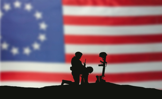 A soldier kneeling in front of the grave of a fallen soldier. American flag in the background. The flag is a Betsy Ross flag of independence.