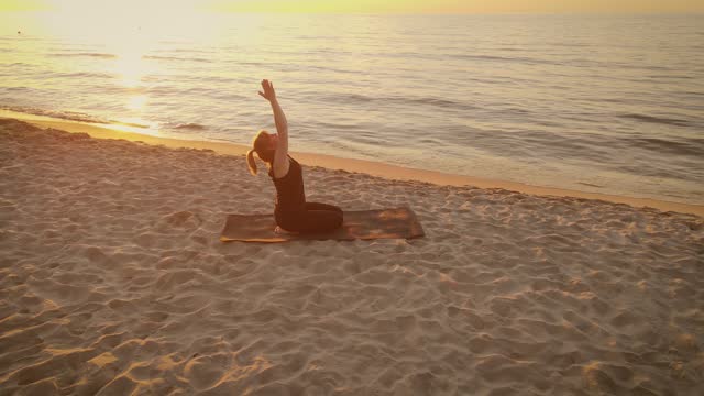 Focused woman practicing meditation raising hands up on sandy beach summer evening. Yoga woman sitting on fitness mat near ocean waves aerial view
