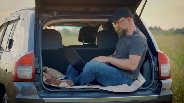 Man working remotely on laptop in nature sitting in car trunk while traveling