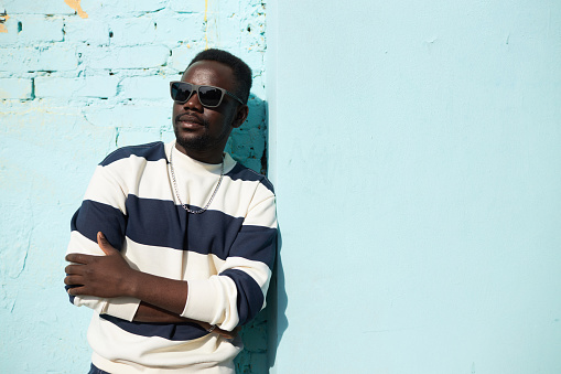 Vibrant waist up portrait of young Black man standing by blue wall in city wearing sunglasses and street style outfit, copy space