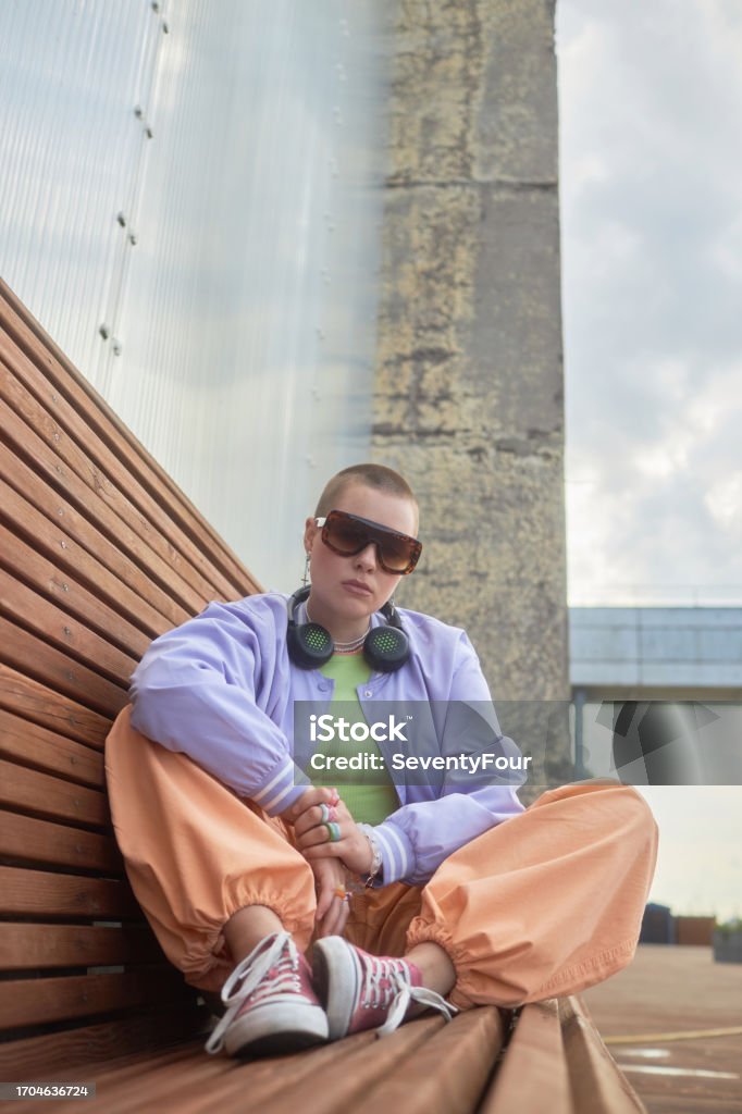 Bald girl sitting on bench in urban city setting and wearing trendy outfit Vertical fashion shot of bald tough girl sitting on bench in urban city setting and wearing trendy outfit 1990-1999 Stock Photo