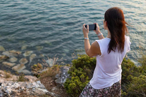 Woman enjoying her summer holiday. Beautiful sea view at summer sunset at golden hour. She is taking pictures with her phone.
