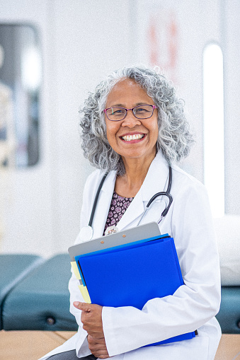 Portrait of a mature adult female physician of Pacific Islander descent smiling confidently at the camera while sitting in a medical examination room.