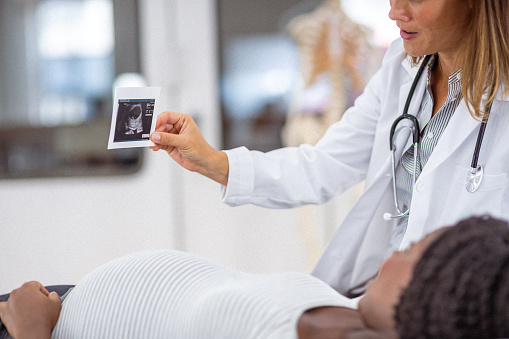 An unrecognizable pregnant black woman lies on a medical examination table while at a medical appointment and looks at an ultrasound scan her female doctor is holding.