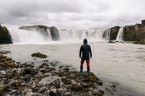 A discoverer is standing next to the river with waterfalls and watching a beautiful view with a dramatic sky in the background. A man is discovering and exploring new places in nature.