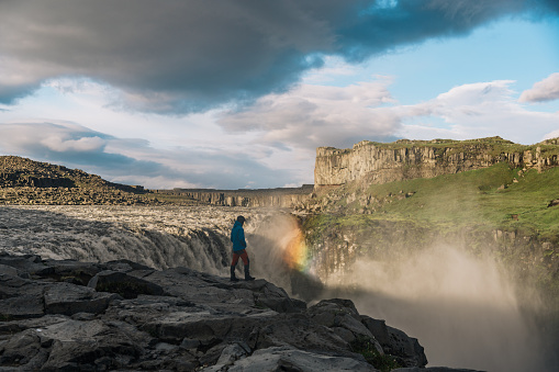 An explorer is discovering beautiful untouched nature in Iceland. A man is discovering hidden places in Iceland. A man is standing next to the waterfall with a rainbow while watching it from a cliff.