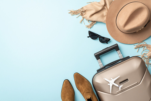 Eleve sus vacaciones de otoño con elegancia. La toma aérea captura una maleta gris, una bufanda suave, botas elegantes y un sombrero de fieltro sobre fondo azul claro, lo que proporciona espacio para su contenido publicitario o textual. photo