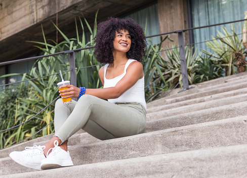 Happy student tourist girl with afro hairstyle sitting in the staircase outdoors. Enjoying an orange juice, resting and taking a break.