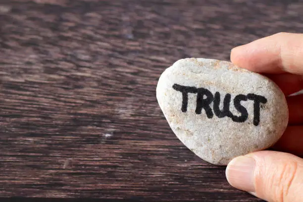 Hand-holding rock with handwritten word "trust" over a wooden background. Close-up, top view. Copy space. Strong Christian faith and belief in God Jesus Christ, biblical concept.