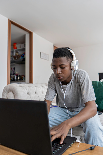 Black Latino teenager studying with laptop at home in living room while listening to music with headphones.