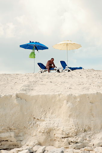 Man reading under a parasol on an edgy beach. An empty chair beside him. Vertical full length outdoors shot with copy space. This was taken in Cayo Largo, Cuba.