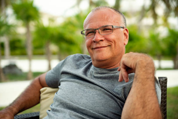 Handsome 50+ man portrait relaxing in a tropical setting. He is wearing a grey top. He is bald, wearing eyeglasses, tanned and smiling at the camera. Horizontal waist up outdoors shot with copy space. This was taken in Cayo Largo, Cuba.