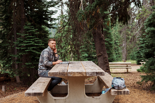 A Caucasian man smiles with gratitude at the opportunity he has to travel and have life balance due to a job working remotely. The man is seated at a picnic table in a forested campground and is using a laptop computer.