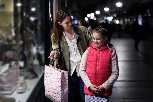 A mother and her daughter create cherished memories as they embark on a nighttime window shopping escapade, their hearts filled with warmth and joy as they admire the captivating displays along the city street, forging a deep bond through shared exploration