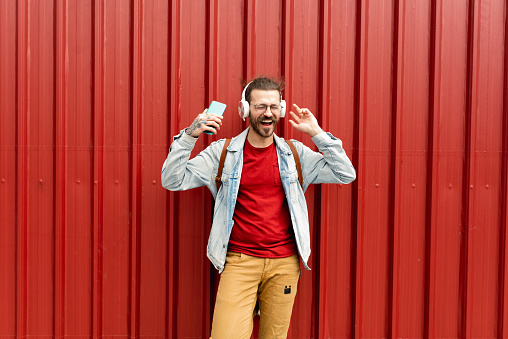 Relaxed and positive man in sweatshirt listening to music in headphones with calm and smiling face isolated on red studio background. Concept of human emotions, casual fashion, lifestyle
