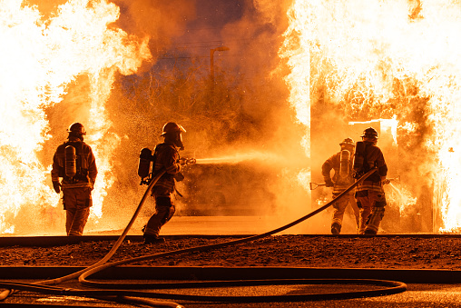 4 firefighters battle a roaring blaze.  2 fire hoses shooting water.