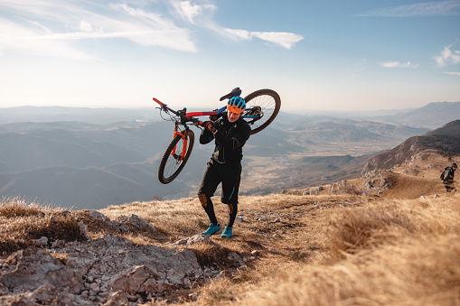 A  lonely tired adult caucasian male carrying his expensive mountain bike on the shoulders and walking uphill in order to reach the top of the mountain. The athlete is wearing all black and a vibrant helmet. Behind the male there is an amazing view of the surrounding valleys and hills. The weather is sunny and warm.