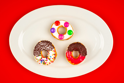 High angle view of doughnuts on white oval plate over red color background. Image made in studio.A doughnut or donut is a type of food made from leavened fried dough. It is popular in many countries and is prepared in various forms as a sweet snack that can be homemade or purchased in bakeries, supermarkets, food stalls, and franchised specialty vendors.