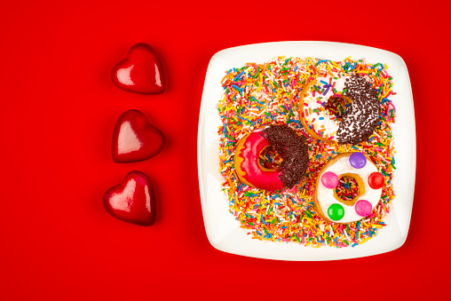 High angle view of doughnuts on white square plate over red color background with sugar sprinkles and heart shape Christmas ornaments. Image made in studio.A doughnut or donut is a type of food made from leavened fried dough. It is popular in many countries and is prepared in various forms as a sweet snack that can be homemade or purchased in bakeries, supermarkets, food stalls, and franchised specialty vendors.