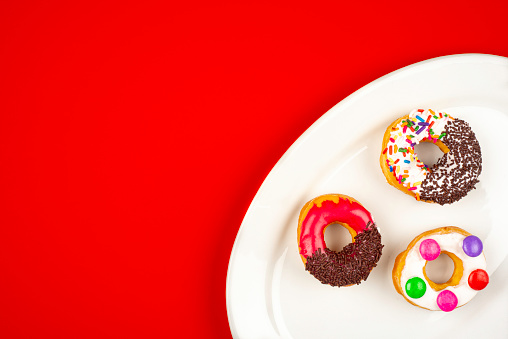 High angle view of doughnuts on white oval plate over red color background. Image made in studio.A doughnut or donut is a type of food made from leavened fried dough. It is popular in many countries and is prepared in various forms as a sweet snack that can be homemade or purchased in bakeries, supermarkets, food stalls, and franchised specialty vendors.