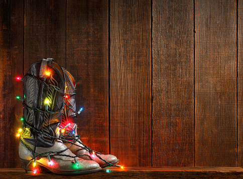 A cowboy boot adorned with Christmas lights sit next to an old wooden fence.