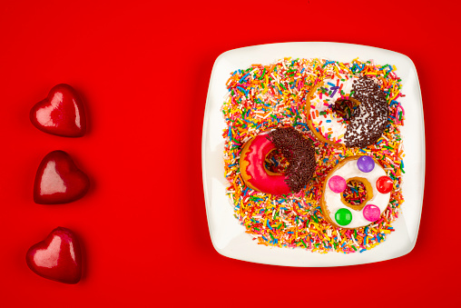 High angle view of doughnuts on white plate with sugar sprinkles over a red color background and three heart shape of plastic.Image made in studio