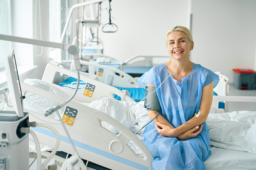 Smiling blonde sits on a special hospital bed, she is connected to a blood pressure monitor and an oxygen machine