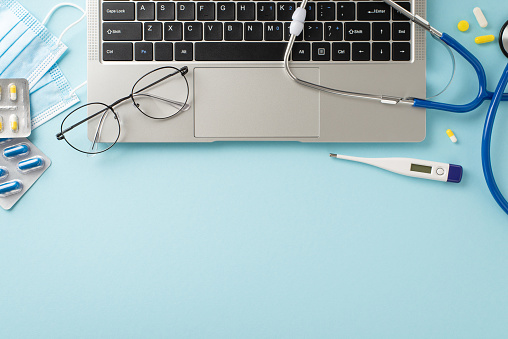 Virtual doctor appointment concept. Top view laptop, stethoscope, glasses, thermometer, pills in blisters, and masks arranged on pastel blue background with space for text or promotion