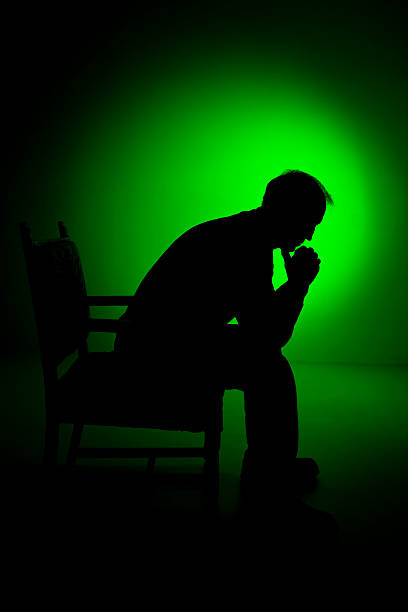 Depressed man in silhouette sitting in a chair.  He has a green spot background behind him.