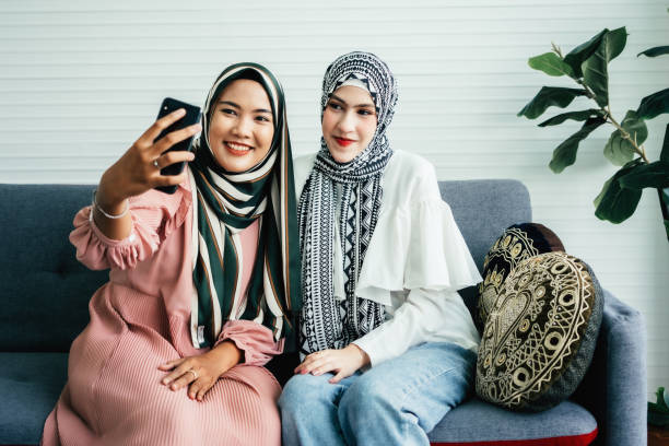 jovens amigas muçulmanas sorrindo enquanto tiram uma selfie juntas - women holding shopping bag living room - fotografias e filmes do acervo