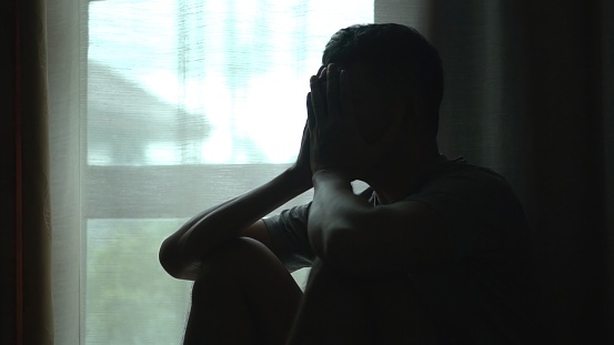 Silhouette of asian man sitting alone by window dark room , head in their hands,  feelings of sadness,depression,hopelessness ,Mental Distress Consept.